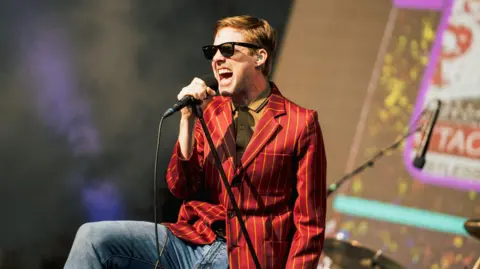 Ricky Wilson of the Kaiser Chiefs, dressed in jeans and a yellow striped red blazer, wearing a yellow shirt and sunglasses, singing into a leaning microphone stand.