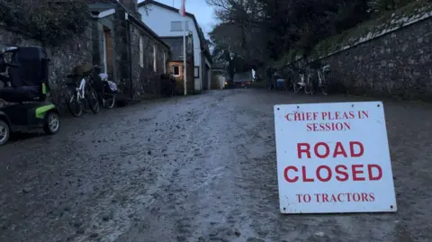 A sign which says Chief Pleas in session, Road Closed to tractors on a muddy road. Along a wall on the left are mobility scooters and bikes. 