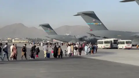 People carrying luggage walk across an airport tarmac towards large US military transport aircraft during an evacuation