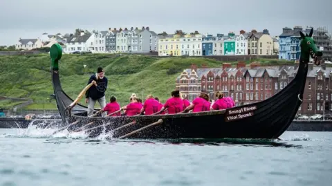 A team of women in pink T-shirts row a boat while the coxswain stands at the stern of the boat holding the rudder. Peel can be seen in the background.
