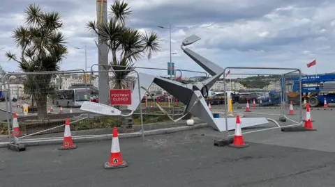 A large steel structure of a triskelion on part of a roundabout ahead of it being mounted. The structure is surrounded by fencing and traffic cones. 