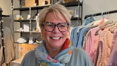 Samantha Gaudin runs a clothes shop in St Aubin. She is wearing glasses and has blonde hair. She is wearing a scarf.
