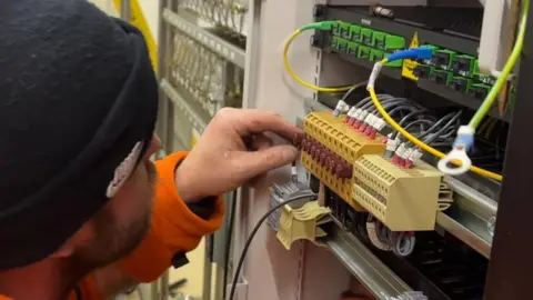 A worker wearing a black beanie and an orange high-vis uniform is working on the wires in a signalling box. 