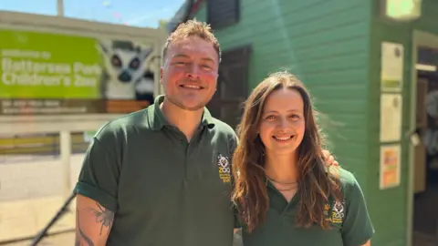 A man with blonde hair and a woman with long brown hair, both wearing green polo shirts, smile into the camera in front of Battersea Park Children's Zoo