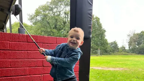 UGC Wilson is mid swing with his new golf club. He is grinning at the camera and holds the driver in the air as he swings it to the right. He has short light brown hair and wears a denim hooded shirt jacket with a grey long sleeve top underneath and black trousers. He is standing in a driving range bay, which has a red brick wall behind him and the green can be seen in the distance, lined by trees.