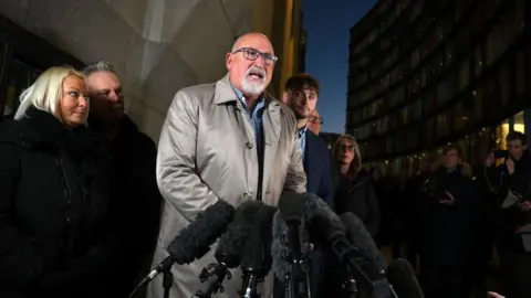 Getty Images Radd Seiger is pictured standing outside a court building before a bank of microphones. He is tall, bald, with a short grey beard and is wearing dark-framed glasses and a beige raincoat over an open-necked blue and white checked shirt. He is flanked by several members of Harry Dunn's family who are looking on.