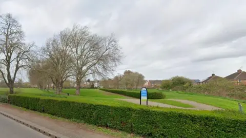 Google Park, with hedge visible in the centre and another hedge at the boundary. There is a blue sign saying "Castle Fields". There are trees to the left, and a road with pavement in the foreground. There are houses in the background.