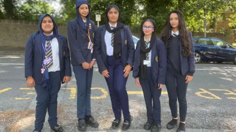 A group of five schoolgirls in dark blue uniforms standing on a roadside next to a yellow 'keep clear' sign