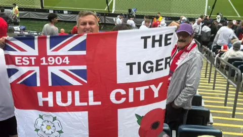 Hull City fan Ed Faulkner holding a customised flag aloft. The flag features references to Hull City on a St George's Cross background. Faulkner is standing in a stadium with other fans around him.
