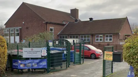 Google Maps A Google Street View image of the entrance to Tudor Church of England Primary School. There are green metal gates and fences on the outside of the school, which is made up of two brown brick buildings. There is a red car parked inside the gates.