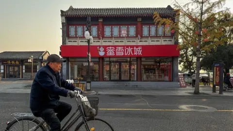 Getty Images An elderly man rides a bicycle past a Mixue Ice Cream & Tea shop on November 4, 2025, in Zhengding, Shijiazhuang, Hebei Province, China. 