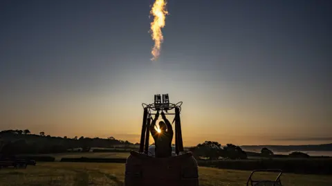 A person standing in a hot air balloon basket fires up the burners against an early morning sky of dark blue and orange