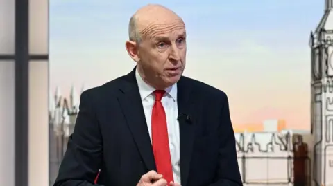 John Healey is wearing a dark suit, white shirt and a red tie.