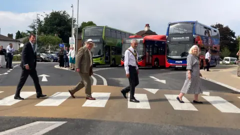 Four people walking on a public crossing, emulating the Beatles album cover. Buses and people in the background.