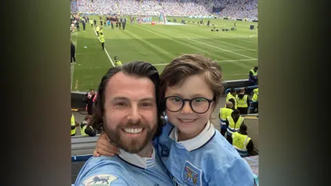 A picture of Matt Bradley on the left smiling at the camera wearing a sky blue Coventry City top and his son on the right smiling at the camera wearing glasses and a sky blue Coventry City top. Behind is the CBS Arena with the green football pitch, fans far in the opposite stands and stewards dotted around.