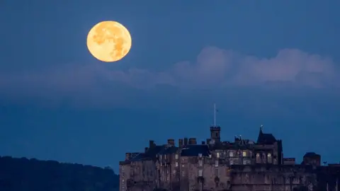 The full October moon, known as the Hunter's sets behind Edinburgh Castle, Scotland. 17 October 2024. The moon is on the left hand side of a blue dusk sky. The castle in in the foreground to the right.