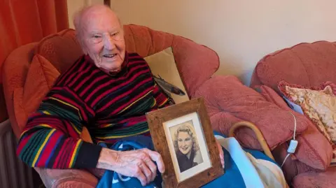 Alex Moss/BBC Aman sitting in an armchair holding a wooden framed picture of a young woman. The man has balding grey hair and is wearing a striped coloured jumper with a blanket over his knees. 