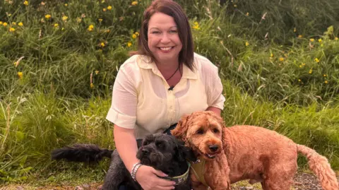 A smiling lady kneels in front of a grassy knoll on which wild flowers are growing. She is smiling and has shoulder length brown hair and is wearing a yellow shirt and black trousers. She is holding two dogs. The dog on the right is sandy coloured and short haired, while the black dog on the left is wearing a blue and yellow collar.