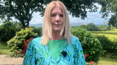 Mel Wallace is standing in her garden on a pebbled section. Behind her are trees, plants and bushes and in the far background is a scenic view of rural Wales with rolling hills and trees visible. Mel is wearing a turquoise and green patterned floaty top which has a button on the chest. Underneath she has on a green t-shirt and a microphone can be seen clipped to her lapel. She has shoulder-length blonde hair and is looking at the camera. It is a head an shoulders shot of her.