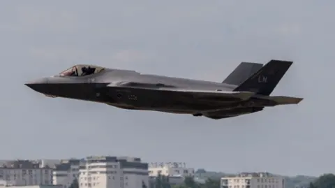 A fighter jet in the air against the sky, with the tops of some buildings visible in the background