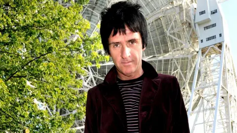 Getty Images Johnny Marr, with a burgundy velvet jacket and black and white striped top, looks into the camera in front of the Lovell Telescope