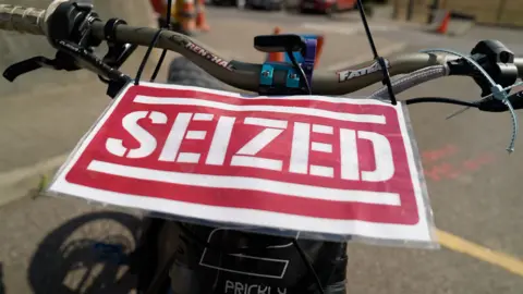 Ben Moore/BBC A red laminated paper sign which reads "seized" in capital letters is attached with cable ties to the handlebars of a bike. In the background there is concrete but not much of the bike is visible other than the brakes and wires and cables.