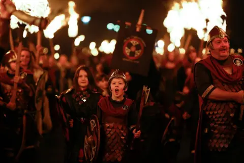 Getty Images A small small boy is dressed like a Viking, carrying a shield and sword. His mouth is wide open. He is at the head of a parade of children and adults dressed similarly. The dark sky is lit up by flames from torches being carried by people following in their wake. 
