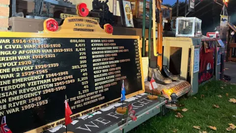 A photo of the Remembrance display at an angle. There is a war board closest to the camera with a list of all the wars and in the background is wooden poppies on the archway that are lit up. There is also a wooden rifle with a war helmet and a set of war boots.