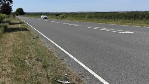 Steve Hubbard/BBC A road where a single white car is travelling toward the camera. The field is lined with bushes, trees and there are fields in the distance. 