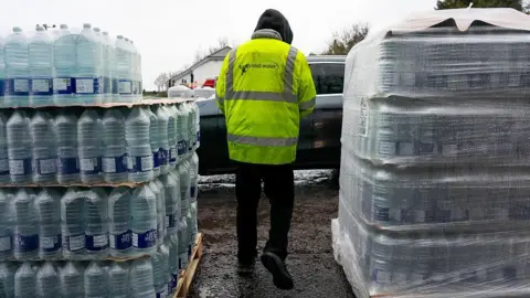 A person in a yellow high vis coat with "South East Water" written on the back. They are walking between two crates of bottled water.