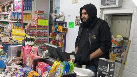 Usman standing in front of a till wearing a black hoodie and gilet with his hands in his pockets. His hood is on his head and he has a black beard. 