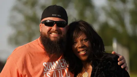 Jon Parker Jon Parker smiles with his arm around a woman. He wears a black cap backwards on his head and an orange T-shirt. The woman has shoulder length black hair and wears a black top.