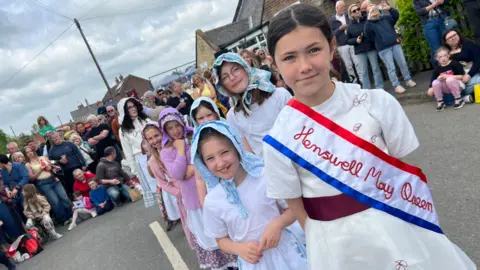 BBC A group of children dressed in traditional parade clothing walk along a village street. The child at the front wears a white outfit with a sash reading "Hemswell May Queen" and is followed by others in white clothing and patterned headscarves. A large crowd of spectators lines the roadside behind them.
