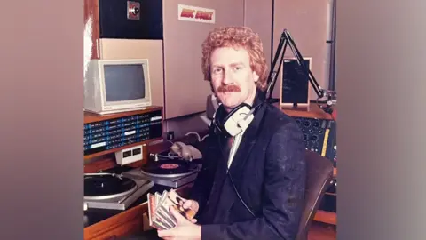 Dave Monk sits in a radio studio with monitors, turntables and mixing desks around him. He has a set of headphones hanging around his neck and he is holding several CDs in his hands. He is wearing a dark jacket with white shirt underneath. He has strawberry-blond curly hair and has a moustache.