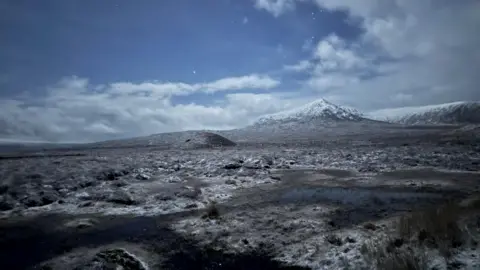 A moonlit winter landscape with a snow‑covered mountain in the distance. The ground is frosty with small pools of water reflecting the sky, and stars are visible through scattered clouds.