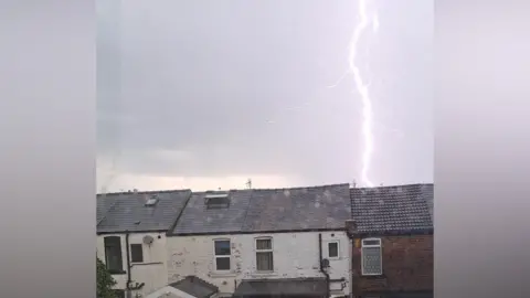 Annika/BBC WeatherWatchers A bold of bright, white lightning reaches down from the sky above a row of terraced houses.