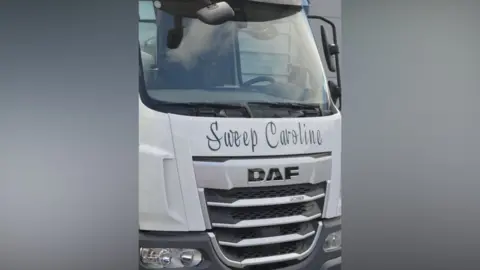 A close-up photograph of the front of what appears to be a white light goods vehicle/lorry. "Sweep Caroline" can be read on the front in black text. 
