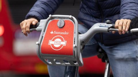 A close up shot of a man wearing a blue puffer jacket riding a Santander e-bike. The photo only shows the handlebar and his hands - no view of his face.