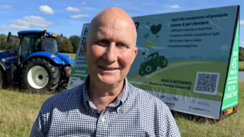 BBC Andrew Gibson standing in front of a blue tractor with a trailer that is advertising prostate cancer figures, the symptoms and urging people to get checked.