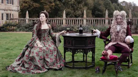 Claire Foy and Richard E Grant starring in Savage House sit on a lawn in front of Montacute House in Somerset wearing period dress.