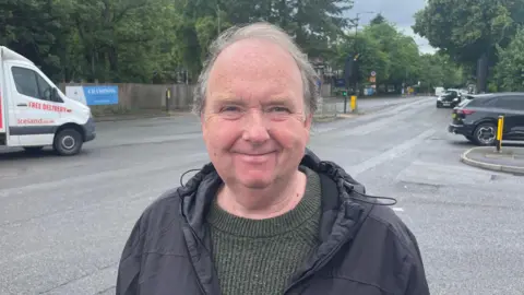 A man wearing a dark waterproof jacket and green jumper stands on a road near a junction. Behind him is a red and white delivery van and a car dealership. The sky is overcast.