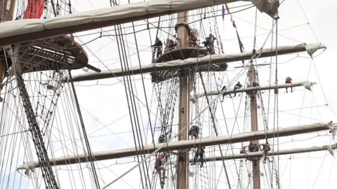 Masts and riggin of a tallship with people wearing harnesses high up in the air, working on the sails