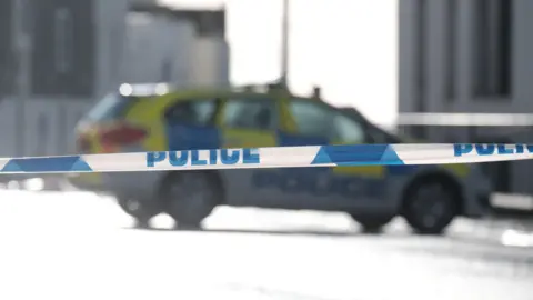  A blurred police car parked on a road in the distance behind police tape.