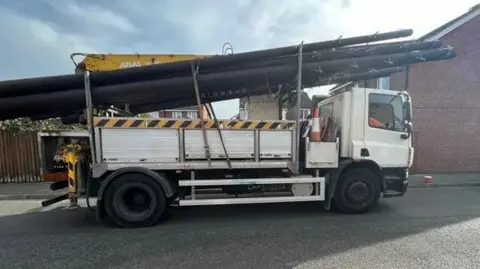 A white lorry carrying half a dozen long brown telegraph poles, which are securely fixed to the back the metal struts, and a yellow crane arm. It is parked on a road on a housing estate.