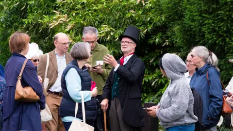 Friends of Foster Hill Road Cemetery Adrian Bean undertaking a talk at the Bedford cemetery. He is surrounded by people and is wearing a Victorian outfit with top hat, jacket and cravat.