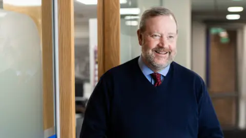 Doug Melville stands in a office and smiles at the camera. He is wearing a jumper, shirt and tie