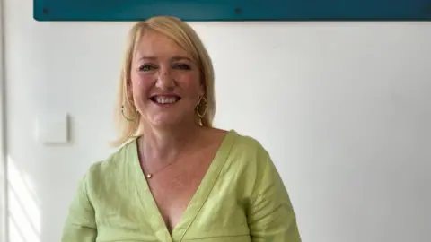 Alongside A woman with short blonde hair wearing a light-green top is posing for a picture smiling. She is standing in front of a white wall.