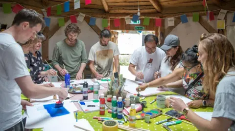 Eden Project Communities A group of nine people gathered round a table painting. There are art supplies piled in the middle of the table