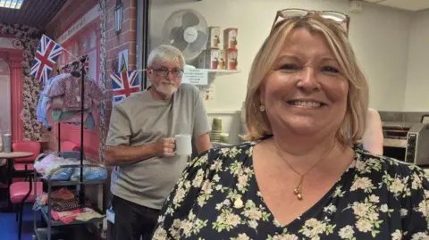 Fiona is smiling and is wearing a black dress with white flowers on it. She has short blond hair in a bob style and is standing in front of a tea and snack window, selling hot drinks and toast. There are Union flags in the background along with a man with grey hair, grey t-shirt and a cup of tea who is looking at the camera.
