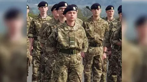 Family Handout Gunner Beck wearing her camouflage military uniform and black cap. She is standing among her fellow soldiers with her arms clasped behind her back and a stern facial expression.